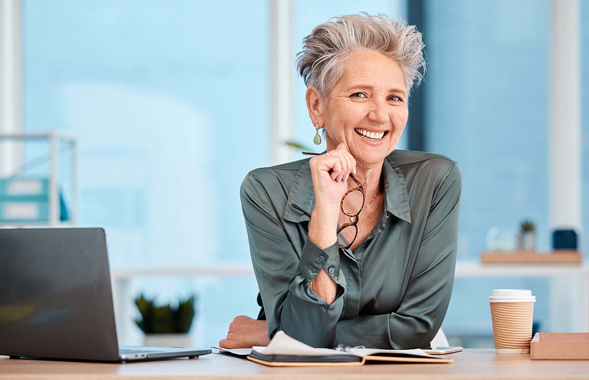 Painless PRP and Low-Level Laser Therapy for Female Hair Loss - Case Study 14 An older female with short grey hair leans on a desk, resting her head on her chin, smiling confidently at the camera.