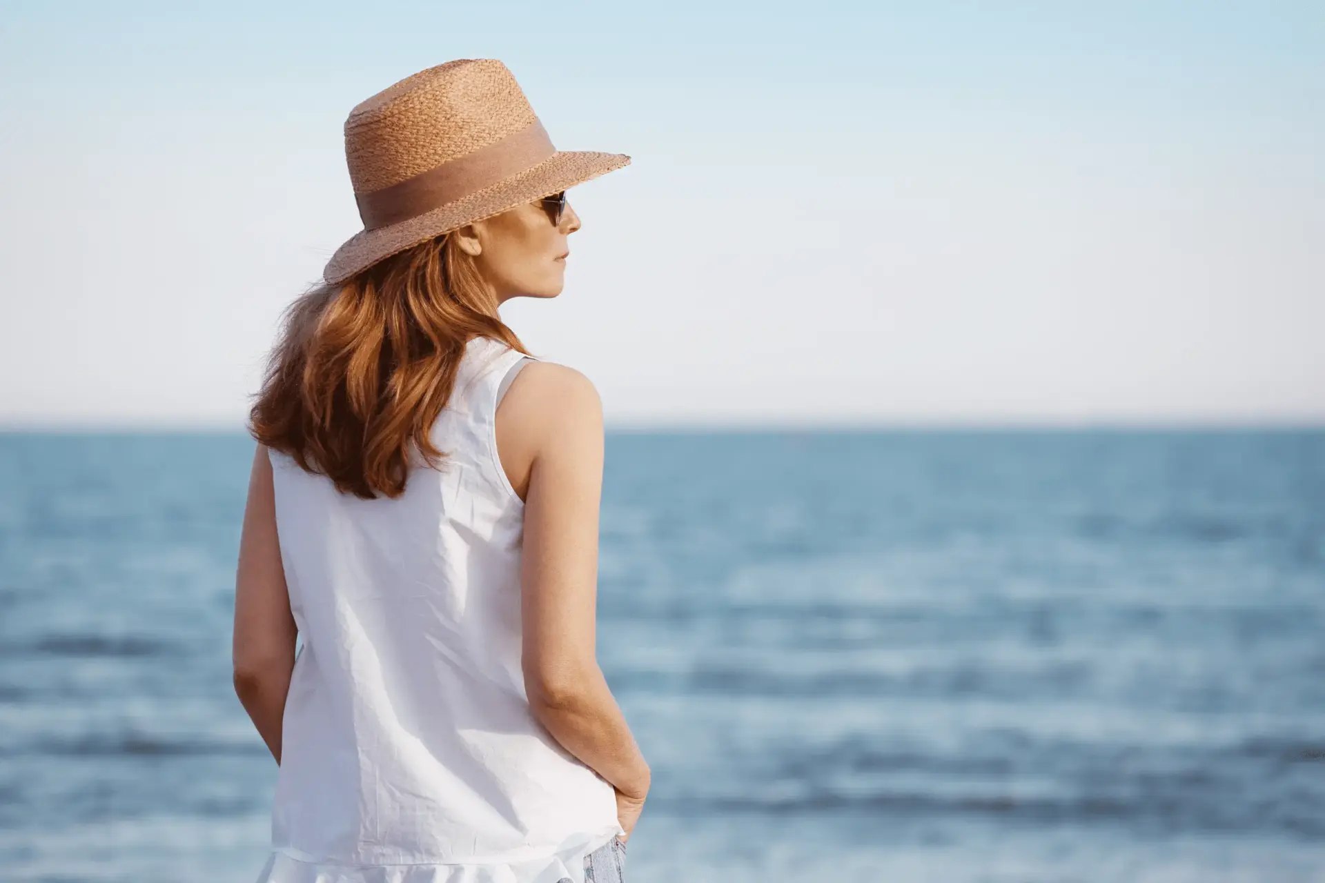 A woman with a white tank top, sunglasses, and a hate looking off into the distance on a beach.